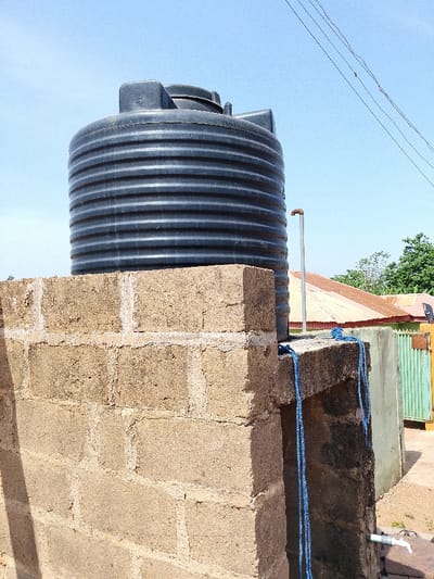 This image depicts an outdoor water storage and dispensing setup in Oke Onigbin, Nigeria, under a clear blue sky. A large, dark grey, cylindrical plastic water tank with horizontal corrugations occupies the upper-center, resting on an elevated platform constructed from light brown/tan concrete blocks with visible white mortar. A silver-colored metal pipe extends vertically from near the tank's base, terminating with a rusty cap. A coiled length of blue rope is draped over the edge of the block platform. Lower down on the concrete structure, a white PVC spigot with associated piping is visible. In the background, part of a building with a rusty corrugated metal roof is visible, along with a section of a green slatted gate or fence and some green foliage. Thin overhead power lines are visible against the sky in the upper right. No individuals are present in the scene.