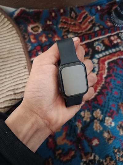 A close-up image depicts a person's left hand holding a black smartwatch against a patterned background. The hand is positioned centrally, displaying the smartwatch with its blank, grey, rectangular screen facing the viewer. The watch features a black frame and a black silicone strap, with two circular buttons visible on its right side. The skin of the hand appears light-toned, with visible veins on the back. A black, ribbed fabric cuff, likely belonging to the person's sleeve, is visible at the bottom left. The background consists primarily of a blue patterned rug or carpet featuring abstract motifs in red, brown, and light cream colors. A light beige, striped object or fabric is partially visible on the far left. The specific location in Denov, Uzbekistan, cannot be determined from the visual evidence present in the image. The scene is indoors under ambient lighting.