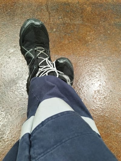 The image shows a close-up view of a person's legs and feet wearing dark blue work pants with reflective stripes and black hiking boots. The boots have white laces and grey accents. The person is standing on a wet, textured surface that appears to be concrete or asphalt, with areas of standing water and what might be salt deposits. The background is blurred, suggesting a shallow depth of field. The scene is likely outdoors, possibly in an industrial or port area, given the practical nature of the clothing and the condition of the ground. The lighting appears to be overcast or diffused. The overall impression is one of a person in work attire, in an environment that is damp and potentially gritty.