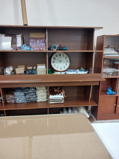 A medium shot, eye-level photograph shows a wooden shelf unit against a white wall. The shelf unit is filled with various items, primarily craft supplies. On the top shelf, there are bundles of beaded trim, fabric, and some small tools, including what appears to be a hot glue gun. A circular clock with a red frame is prominently displayed on the second shelf from the top. This shelf also holds several small white bowls containing what look like beads or sequins, and some other assorted craft items. The third shelf from the top contains more bundles of beaded trim and some packaged beads. The bottom shelf is less visible, but appears to hold more packaged craft supplies. To the right of the main shelf unit is a smaller, glass-fronted cabinet, also containing shelves with various items, some of which appear to be fabric or rolls of material. A bright blue plastic bag is visible on one of the shelves in this cabinet. In the foreground, a portion of a light brown, reflective surface, possibly a table or countertop, occupies the bottom of the frame. The overall impression is of a craft room or workshop. The location is specified as being in the City of Türkmenabat, Turkmenistan.