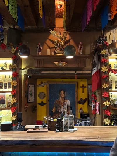 Mexican restaurant interior features colorful papel picado hanging from the ceiling. A Mexican flag is draped above a display of liquor bottles with poinsettia garlands. A painting and various religious artifacts adorn the wall, illuminated by two spherical light fixtures. A wooden bar dominates the right side of the frame.