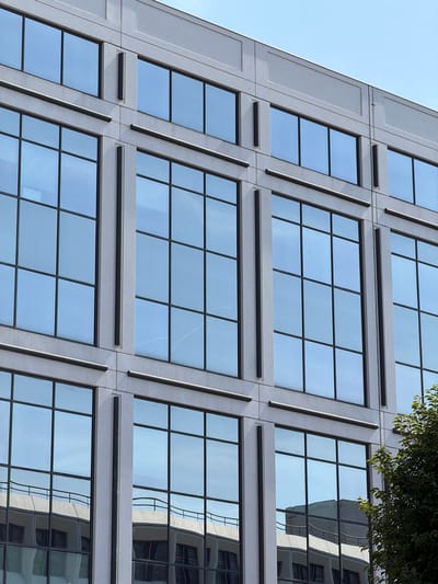 The image shows a close-up, low-angle view of the exterior of a modern office building, located in the city of Rungis, France. The building features a facade of concrete panels and large, grid-like windows with reflective blue glass. The windows are arranged in a repeating pattern of rows and columns. There are dark vertical accents between some of the window panes and horizontal dark bands above some rows of windows. In the lower right of the image, a portion of a green leafy tree is visible, and the reflection of another building is visible in the lower windows. The sky is clear and blue, indicating a daytime scene. There are no people or animals visible in the image.