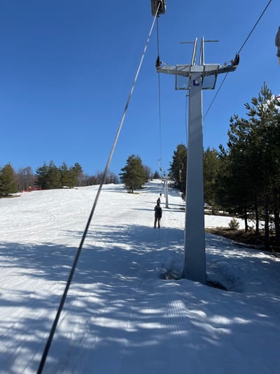 The scene is situated on a snow-covered ski slope in Bogoslov, Bulgaria, under a clear blue sky. The perspective is from a ski lift, evidenced by a prominent dark cable extending diagonally from the upper left foreground. A tall, light-grey metal ski lift support tower with a lattice platform and pulley system stands on the right side of the frame. On the groomed snow below, a person wearing dark trousers and a dark and red-toned jacket walks uphill, away from the viewer. A second, smaller figure is visible further up the slope, also ascending. Multiple additional ski lift towers are linearly arranged uphill in the distance. The ski slope is flanked by evergreen trees, with some bare deciduous trees visible on a distant hill to the left. Long shadows are cast across the snow, predominantly on the right side of the slope.