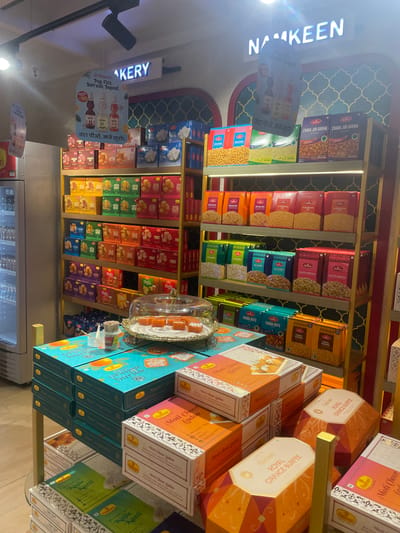 The image depicts a retail display of Indian sweets and snacks, likely in a store in Gollapalle Khurd, India. The scene is well-lit and organized, showcasing a variety of packaged food items on shelves. Prominently displayed are boxes of namkeen (savory snacks) and sweets, with brands like Haldiram's visible. The packaging features vibrant colors and intricate designs, typical of Indian confectionery. Some items, like "Chana Jor Garam" and "Moti Choor Ladoo," are clearly labeled. In the foreground, a clear glass cloche covers a selection of orange-colored sweets, possibly barfi or ladoo, arranged on a decorative platter. Adjacent to this display are stacked boxes of "Nagpur's Orange Burfee" and other sweet boxes, including "Kaju Katli." The overall environment suggests a food retail setting, possibly a sweet shop or a grocery store section dedicated to Indian snacks and sweets. There are no people visible in the image.