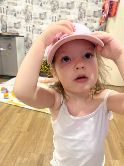 The media file shows a close-up, rotated view of a young child. The child is wearing a white tank top and holding a pink hat on their head with their hands. Their eyes are wide open, and their mouth is slightly agape, suggesting curiosity or a playful expression. The background is blurry, but it appears to be an indoor setting with patterned wallpaper and a wooden floor. A child's play mat with colorful shapes is visible on the floor. The lighting suggests it is daytime. The child's skin is fair, and they have light brown hair. The focus is primarily on the child's face and upper body.