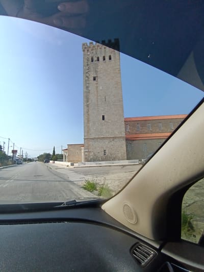 The image, taken from inside a vehicle, shows a prominent stone bell tower and an adjacent building with a red tiled roof. The bell tower is tall and rectangular with crenellations at the top and several rectangular windows along its height. The building next to it has a series of arched windows. The scene appears to be in an urban or semi-urban setting, with a road and other structures visible in the background. The sky is clear and blue. The location is identified as the Municipal Unit of Xylokastro, Greece, likely a religious or historical site given the architecture. The perspective suggests the vehicle is driving past the location.