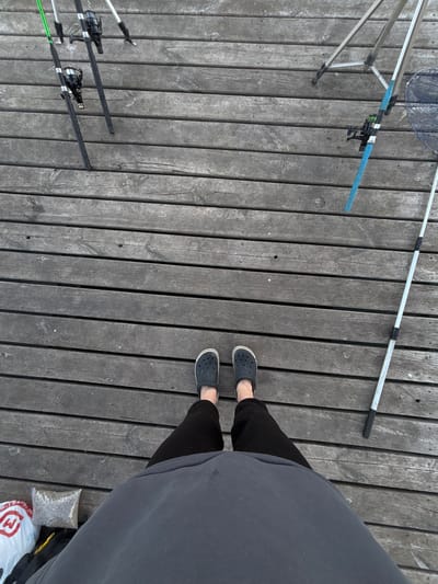 The image depicts a person standing on a wooden dock, possibly a pier or walkway, near a body of water. The person is wearing dark-colored Crocs and dark pants, with a large grey sweatshirt covering much of their torso. Fishing equipment is scattered around the dock, including fishing rods, a net, and a rod holder. There's also a bag of bait or feed near the person's feet, and a shopping bag. The location is unknown; no identifiable details link it to Khryashchevka, Russia.