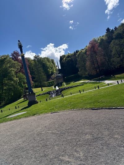 The image shows a park scene in Cleves, Germany, under a blue sky with scattered clouds. A statue stands on a tall column in the foreground. Adjacent to a gravel path is a green lawn. A fountain, trees, and a distant structure are visible in the background.