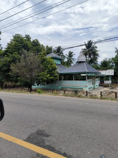 This media file captures a daytime roadside view from a moving vehicle in Sawang, Indonesia. The primary subject is a distinct light blue and white building, characterized by its dark grey corrugated metal roof and a prominent, textured white dome-like cupola at its apex, possibly indicative of a mosque or a traditional Indonesian structure. The building features an elevated veranda with intricate white railings and decorative valances along its eaves. A smaller, simpler structure with a similar roof is partially visible to the right and behind the main building.
The buildings are nestled amidst a dense canopy of lush green trees, including several tall palm trees. Power lines crisscross the partly cloudy sky above. In the foreground, an asphalt road with a solid yellow line on the left and a faint white line in the distance indicates movement. The verge between the road and the building is rough, with rocks and scattered vegetation. A glimpse of a car's side mirror in the lower-left corner confirms the perspective. The bright yet cloudy conditions suggest it is daytime, likely late morning or early afternoon. No people or visible text are present in the image.