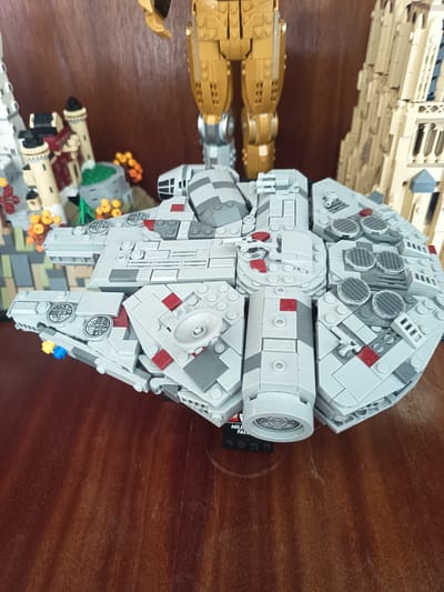 A LEGO model of the Millennium Falcon is displayed on a wooden surface. The model is primarily gray with red accents and features intricate details such as cannons, engines, and circular vents. In the background, other LEGO constructions are visible, including a tall, beige, gothic-style building and a gold robot figure. The wooden surface appears to be a table or shelf. The image does not contain any people or depict the town of Millstreet, Ireland.