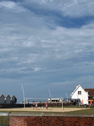 The image depicts a sunny day at a beachside location, likely in Hautot-sur-Mer, France, indicated by the architecture of the buildings and beach huts. The scene is oriented vertically. In the foreground, a sandy area suggests a beach or sports court, with several people engaged in activities. Some individuals are playing volleyball on a court marked by a net and poles, while others are gathered nearby. In the background, a white building with a red-tiled roof stands prominently, possibly a cafe or beach club, with people visible on its terrace. Further back, a row of small, grey beach huts with white trim lines the edge of the sandy area. The overall atmosphere appears to be recreational and lively. The weather is fair with a bright blue sky partially obscured by scattered, wispy white clouds. The lighting suggests it is daytime, likely late morning or early afternoon. The brick facade of a building is visible on the right side of the image, adding a structural element to the coastal setting.