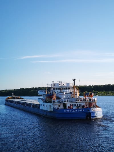 An overhead perspective shows a blue and white cargo barge, identified by the Cyrillic text "ВОЛГО-ДОН 5069" on its stern, moving through dark blue water under a clear blue sky. The barge is long and flat-topped, loaded with yellow cargo. The stern of the barge features a white superstructure with several windows, railings, and lifeboats. A dense green forest lines the distant bank of the waterway. The image is taken in the city of Sud'bitsy, Russia.