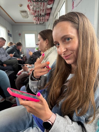 The image captures a selfie-style shot of a young woman inside what appears to be a passenger ferry or boat. The main subject, with long, light brown hair and blue eyes, looks directly at the camera with a slight smile. She is wearing a light-wash denim jacket over a grey hoodie. Her right hand, visible in the foreground, features bright pink and orange nail polish, and she wears a small gold ring. Her left hand holds a vibrant pink smartphone, and a pink smartwatch with a black face is visible on her left wrist.

The setting is an interior space with rows of seats, suggesting a public transport vehicle. Several other passengers are seated in the background, mostly out of focus. They appear to be engaged in their own activities, some looking at phones. Natural light streams in through windows along the side of the vessel, indicating daytime. On the upper left side of the frame, metal racks holding items in clear plastic bags are visible, possibly supplies or cargo. A small, blurry sign with a red border and white background, featuring illegible text (possibly Cyrillic), is visible on the wall in the distance. The overall scene suggests a routine journey for the passengers. The specific location, Nizhneleninskoe, Russia, is provided, but no explicit visual cues within the image confirm this.