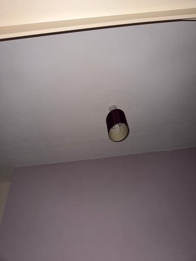 The image shows an indoor scene, likely a residential interior, with a light fixture mounted on a wall. The fixture has a burgundy shade, cylindrical in shape, and appears to be a sconce type. A lightbulb is visible within the shade. The wall it's mounted on is textured and painted in a light greyish-white hue. Adjacent to this wall is another one painted a darker shade of greyish-purple. The corner where the two walls meet is also visible. To the left of the frame is a portion of a door frame, painted white. No people or other objects of significance are present. The image does not provide enough information to determine the specific location within Cwmbrân, United Kingdom.