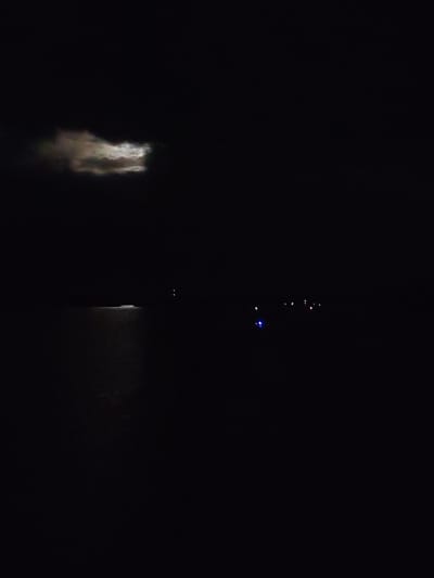 The image presents a very dark, high-contrast nocturnal scene over a body of water, likely a lake or river in Kurikaevo, Russia. The dominant feature is the pervasive blackness of the night sky and water.
In the upper left portion of the frame, a segment of the night sky reveals a distinct cloud formation. This cloud is partially illuminated, likely by a moon obscured behind it, creating varying shades of gray and white and giving it a textured, ethereal appearance against the dark sky.
Below the sky, a vast expanse of dark water fills the lower half of the image. On the left side of this water body, a bright, elongated strip of light is reflected on the surface, indicating an unseen light source just outside the frame or very close to the left edge. Further to the right, across the dark water, several small, distant artificial lights punctuate the darkness. A prominent blue light stands out, accompanied by a few fainter red and orange points of light, suggesting the presence of distant boats, buoys, or a remote shoreline.
There are no discernible people or specific objects other than the distant lights and the illuminated cloud. No activity is taking place, and the scene conveys a profound sense of nighttime stillness and solitude. No text is visible in the image.