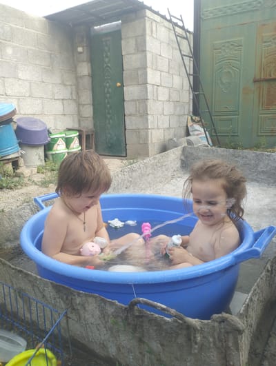 Two young children, a boy and a girl, are having a bath in a large blue plastic tub filled with water. The tub is situated in an outdoor courtyard. The boy, on the left, is partially submerged and holding a pink toy. The girl, on the right, is also in the tub, smiling and with water spraying in front of her. She is holding a white bottle. Both children appear to be enjoying themselves.
The setting is rustic, with a brick wall in the background and a green metal door with decorative panels. A ladder leans against the wall next to the door. Several plastic buckets, including blue and purple ones, are stacked to the left of the wall. There are also green barrels with some visible text. The ground is uneven and appears to be dirt and concrete. The weather seems to be fair, possibly a sunny day given the lighting. The overall impression is one of a simple, everyday moment of childhood fun.