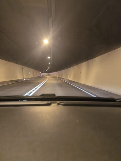 The image shows a view from the driver's perspective inside a car, looking forward through a windshield at the entrance to a tunnel. The tunnel interior is illuminated by overhead lights and lights embedded in the walls. The road surface is asphalt, marked with white lane dividers. The tunnel curves to the right and appears to be for vehicular traffic. The sky above the tunnel entrance is visible and appears overcast. There are no visible people or animals in the image. The scene depicts a common infrastructure element within a city, likely used for transportation. The image itself does not contain specific geographical markers to pinpoint its location within Hornberg, Germany.