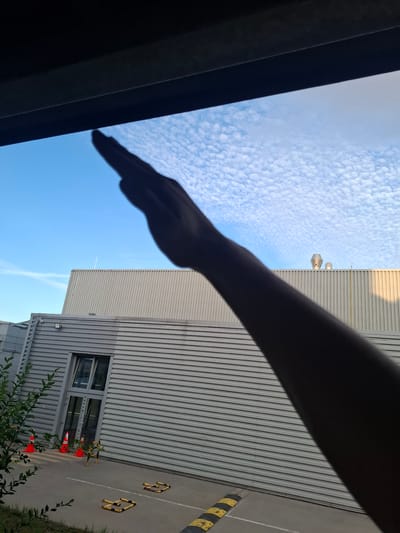The image, taken from a low angle, shows the silhouette of a person's arm and hand extended upwards against a bright blue sky filled with scattered, puffy white clouds. The hand is held flat, with fingers together, as if making a gesture. Below the sky, a corrugated metal building is visible, with several industrial-looking vents or structures on its roof. In the foreground, there are orange traffic cones and what appear to be speed bumps on an asphalt surface. The overall scene suggests an outdoor, possibly industrial or commercial area, bathed in daylight. The image was captured in Sedlec, Czechia.