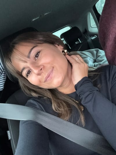 A female individual is seated in the front passenger seat of a vehicle in Valence, France. She has medium-length brown hair with lighter highlights and bangs, and is looking directly at the viewer with a slight smile. She wears a dark, long-sleeved top, a gold-colored circular pendant necklace, and prominent gold-colored hoop earrings. A silver ring is visible on the ring finger of her left hand, which is raised and positioned near her right ear. A grey seatbelt is securely fastened across her chest and lap, displaying an "AIRBAG" label on the seat fabric to her left. The vehicle's interior includes a black fabric seat and a black headrest. In the background, beyond the seat, blurred forms suggest a light grey blanket or garment and a dark red or maroon textured object. The exterior visible through the vehicle's window appears overcast and indistinct.