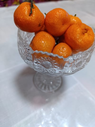 The image, captured in the city of Asaka, Uzbekistan, displays a clear, cut-glass fruit bowl filled with tangerines. The bowl, featuring intricate etched geometric patterns and a short pedestal base, holds approximately eight to ten bright orange tangerines or mandarins. One tangerine at the upper left retains a small green stem. The vessel rests on a white surface, possibly a tablecloth, which exhibits subtle wrinkles and diffuse reflections from an overhead light source. The background is softly blurred, showing indeterminate warm-toned colors. No individuals or specific actions are depicted; the scene is a static indoor arrangement.
