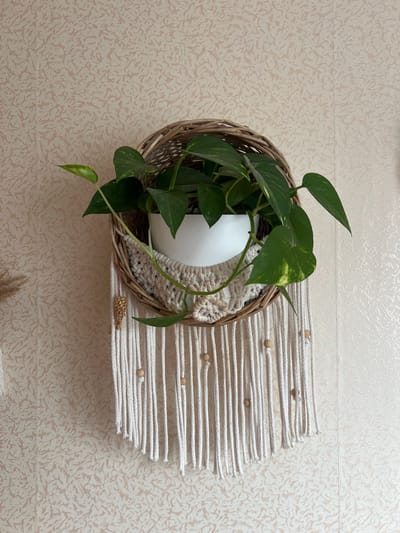 The image shows a macrame plant hanger with a white pot containing a green pothos plant. The hanger is made of white rope and decorated with wooden beads. A small, woven ornament hangs from the side of the basket. The plant hanger is attached to a wall with textured, light-colored wallpaper. The background is an indoor wall. The image does not contain any information about the city of Yakshur-Bod'ya, Russia.