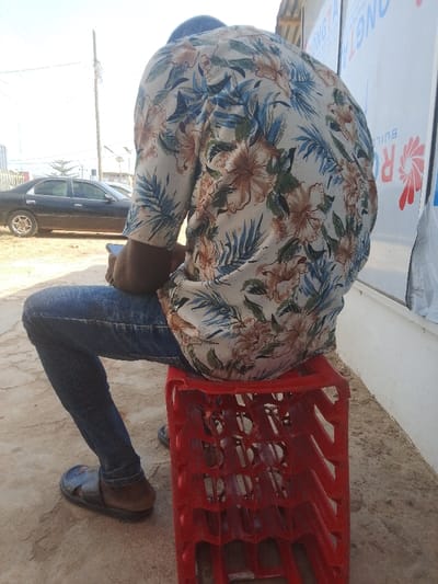 An individual, observed from the back, is seated on an inverted red plastic bottle crate in Abeere, Nigeria. The person wears a light-colored, short-sleeved shirt patterned with blue leaves and brown floral elements, blue denim jeans, and dark sandals. The right hand holds a dark-colored smartphone. The ground surface consists of dry, light-colored earth. In the left background, a black sedan is parked beneath a utility pole. To the right, a white building facade displays a partial sign with blue text "ONGTA BUILD" and a red and white logo.