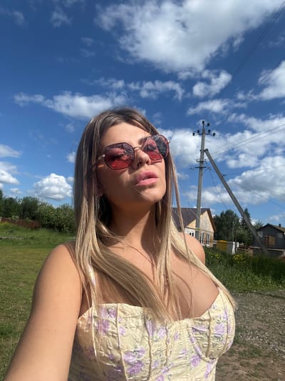 A fair-skinned woman with blonde hair and pink sunglasses is captured in a low-angle, outdoor selfie. She is wearing a light yellow corset-style top with a floral pattern in shades of purple and pink. The background features a bright blue sky with scattered white clouds, a utility pole with wires, some trees and foliage, and a few buildings, suggesting a rural or semi-rural setting. The overall impression is one of a sunny day in a village. Based on the provided information, this scene is located in the village of Pastukhovo, Russia.