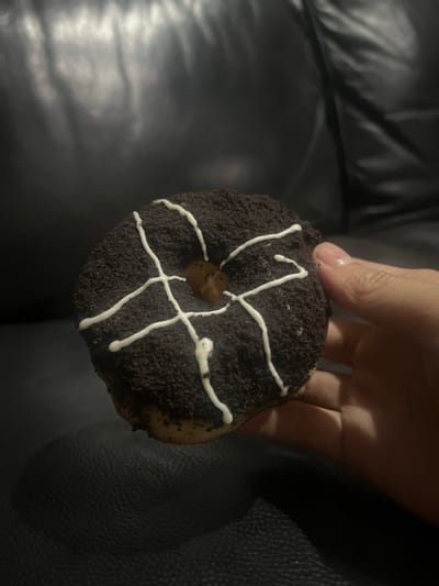 A person's hand is holding a chocolate-covered donut with white icing drizzle. The donut has a hole in the center and appears to be coated in crushed chocolate cookies. The background is a dark, textured surface, possibly a leather couch. The scene is dimly lit, with the light source appearing to be from above and to the left. The location is the city of Kalijaten, Indonesia.