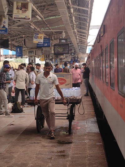 The image depicts a busy railway platform at Itarsi, India, with a passenger train on the right side. A man in a white polo shirt is pushing a cart loaded with bottled water. He is wearing light brown pants and sandals. The platform is crowded with people, many of whom are waiting or moving around. Signs in Hindi are visible on the overhead structure, indicating platform numbers and advertisements. The environment appears to be an outdoor train station with a covered platform roof.