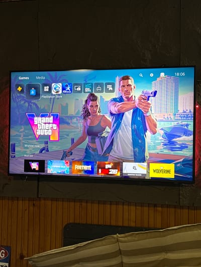 The image shows a television screen displaying the home screen of a Playstation 5 console. The background theme appears to be from Grand Theft Auto: Vice City, featuring characters and imagery associated with the game. Several game icons are visible, including Grand Theft Auto, Fortnite, and NBA 2K26. Other app icons for PlayStation Store, media apps, and system settings are also present. The time displayed on the screen is 18:06. The television is mounted on a wall with a stone-like texture. A portion of a striped couch is visible on the right side of the image. The image provides no indication of being taken in Lilkovo, Bulgaria.