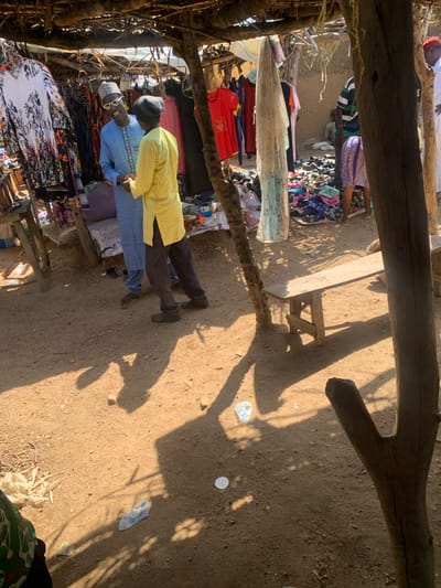 The image depicts an outdoor market scene in Dull A, Nigeria, bathed in the bright sunlight of what appears to be midday. The foreground is dominated by rough-hewn wooden structures, likely part of a stall or awning, casting long shadows across the dusty ground.

The central subjects are two men standing near a collection of colorful textiles and clothing. The man on the right is wearing a bright yellow robe and a dark beanie, with his head tilted slightly downwards. To his left, a man in a blue traditional outfit and a patterned cap is looking towards the camera, wearing sunglasses. His expression appears neutral or perhaps slightly amused.

Behind the men, various garments are displayed, including patterned dresses and other clothing items hanging on racks or folded. The ground is dry and sandy, with some scattered debris, including a few plastic wrappers and a small white object. In the background, more market stalls and goods are visible, suggesting a bustling commercial environment. The overall impression is of a vibrant, casual market day. There is no visible text in the image.