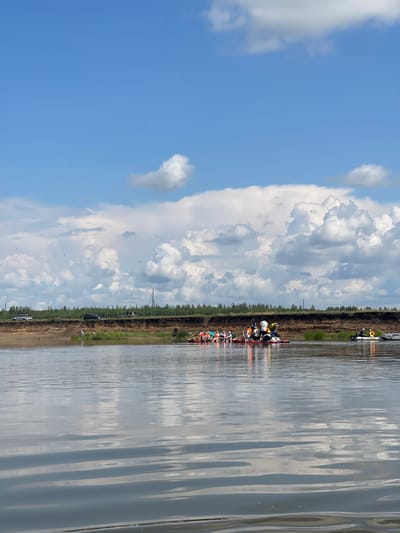 In a wide, calm river under a partly cloudy sky, a group of people are on a raft in the water. The raft is made of several inflatable kayaks and paddleboards tied together. Approximately ten people are visible on the raft, some standing and some sitting. They are wearing casual clothing, with some individuals wearing orange or yellow life vests. The riverbank on the far side is visible, with a brown, eroded cliff face topped by a line of green trees. A few vehicles are parked on top of the cliff. To the right of the raft, two smaller inflatable boats are also on the water. The scene appears to be a recreational outing on a river, possibly near Nezhinka, Russia, given the typical landscape and activity.