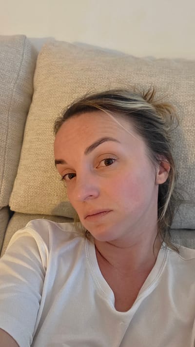 A young woman with blonde hair pulled back in a bun is lying on a light-colored couch. She is wearing a white t-shirt. The woman's face is turned towards the camera, and she has a neutral expression. The background is the textured fabric of the couch and a hint of a wall. The image appears to be taken indoors, likely in a residential setting in Guía de Isora, Spain.