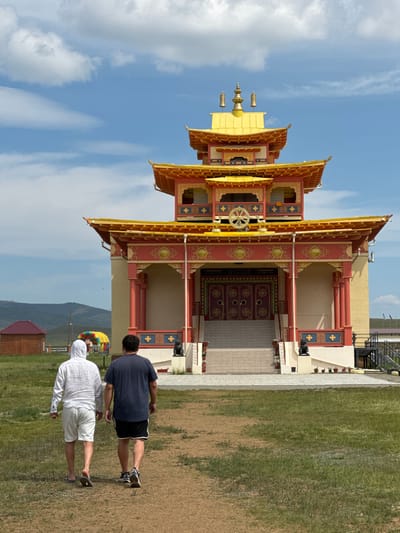 Two men walk away from the camera on a dirt path towards a colorful Buddhist temple in Orongoi, Russia. The temple has a multi-tiered roof with golden accents and red and yellow decorations. The man on the left wears a white hooded sweatshirt and white shorts with sandals. The man on the right wears a dark blue t-shirt and black shorts with sneakers. In the background, there are rolling hills, a small wooden building, and a colorful inflatable structure. The sky is blue with scattered white clouds.