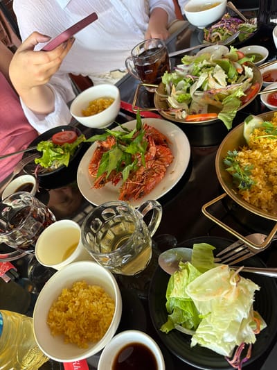 The overhead, close-up shot shows a table laden with a meal, likely in a restaurant, on a sunny day in Tuy Hoa Ward, Vietnam. A person, wearing a white shirt and pink pants, is in the upper left, holding a maroon phone. The table is black and features several dishes: a plate of grilled shrimp with greens, a bowl of yellow rice, a salad with lettuce and shredded vegetables, a bowl of rice, and a dish with fried rice. Drinks in clear glasses and small bowls of sauce are also present. The overall scene depicts a shared meal.