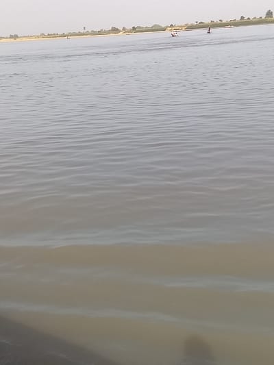 The image depicts a wide river in the city of Ibi, Nigeria, under a pale, diffuse sky. The water surface, appearing murky grey-brown, exhibits numerous small ripples. In the mid-distance, a white boat, consistent with a canoe, is observed slightly to the left, carrying at least one individual. Further right, a distinct red object, possibly a small boat or a standing person on a watercraft, is visible afloat. A third, smaller, dark object or figure is present closer to the distant shoreline on the left. The distant shoreline itself features a low, sandy bank sparsely vegetated with low green growth and a singular, taller tree. The immediate foreground consists solely of the river's surface, with a dark, indistinct shadow present at the lower-left corner.