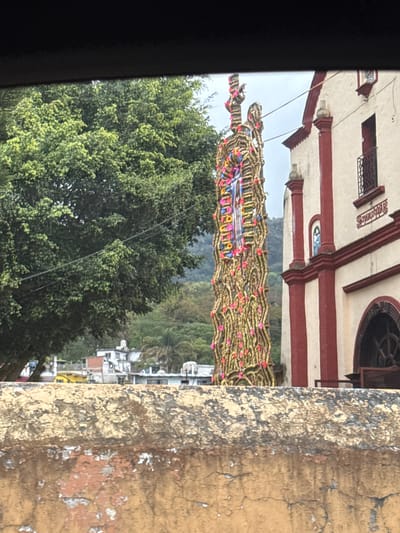 Captured through a vehicle window, the image presents a scene in San Pablo Coapan, Mexico. The foreground shows a portion of a deteriorated, weathered wall with peeling paint, characterized by a range of yellow hues and visible decay. The background transitions into a lush expanse of trees, obscuring the skyline. Centrally positioned is a wooden figure adorned with extensive floral arrangements in yellow, white, and pink, possibly representing a saint or religious figure. This arrangement is suspended above a public street, as indicated by the presence of residential buildings and possibly a church. The architecture features red and white horizontal striping. The overall composition suggests the image was captured during a public event or religious procession.