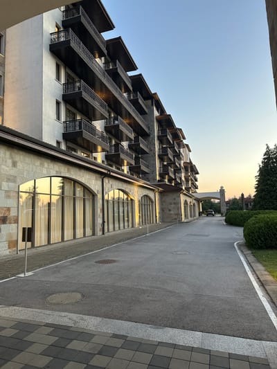 The image depicts the exterior of a hotel building in Razliv, Bulgaria, at dusk. The hotel has multiple stories with dark balconies and a stone facade on the lower level. Large arched windows with sheer curtains are visible on the ground floor, reflecting the warm glow of the setting sun and trees. A paved driveway leads towards the hotel, curving to the right. A dark SUV is parked on the driveway near an arched gateway. Green hedges and trees flank the driveway and hotel. The sky is a clear, soft blue with a gradient of orange and yellow towards the horizon, indicating sunset. The foreground shows a patterned paved walkway. There are no people visible in the image.