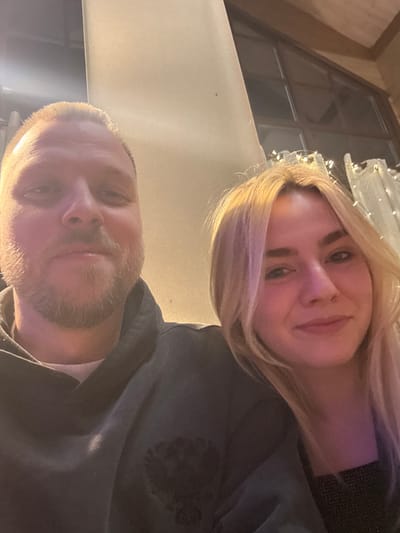 A selfie captures a woman with blonde hair and fair skin, positioned above a man with a bald head and a beard. Both are looking at the camera. The woman wears a dark top, and the man is in a dark sweatshirt with a light undershirt visible at the collar. Behind them, a window with visible panes is adorned with string lights. The setting appears to be indoors.