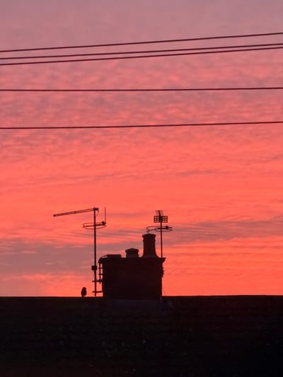 The scene, set in Bretton, United Kingdom, displays a silhouetted urban rooftop against a sky characterized by intense red and orange hues at the horizon, transitioning to lighter pinks and purples with textured cloud formations overhead. Four dark, parallel utility lines are visible horizontally across the upper sky. On the dark, silhouetted rooftop, a single small bird is perched on the left side, adjacent to a prominent chimney stack. To the left of the chimney, a pole supports a Yagi-style television antenna and a circular antenna. To the right of the chimney, another pole holds a grid-like antenna above a circular antenna.