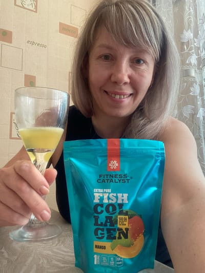 In the city of Kursk, Russia, a Caucasian woman with shoulder-length blonde hair and a fair complexion smiles at the camera. She is holding a glass filled with a yellow liquid and a turquoise package of "FISH COLLAGEN" supplement. The background features a tiled wall with various coffee-related images and text, including "espresso" and "mocco". The woman is wearing a dark top, and the scene appears to be indoors.