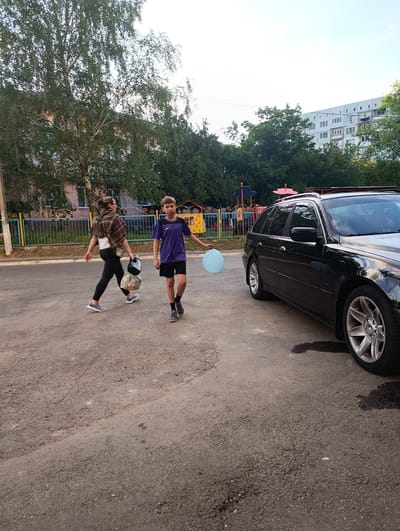 A young boy in a purple t-shirt and black shorts, holding a light blue balloon, walks on an asphalt road in front of a black station wagon. To his left, a woman in dark leggings, a white shirt, and a brown plaid shawl walks away from the camera, carrying two bags. A colorful playground fence is visible behind them, with trees and a multi-story building in the background. The scene appears to be in an urban residential area in Rezina, Moldova.