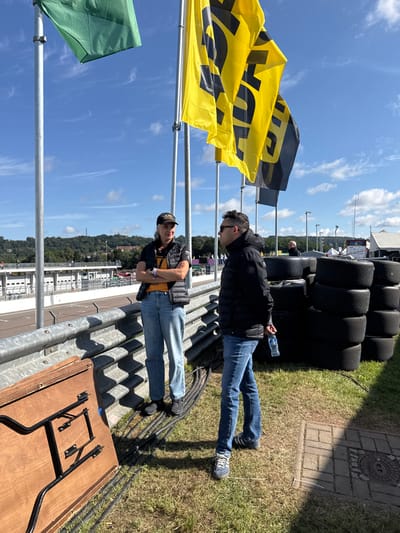 The image depicts an outdoor scene at a motor racing circuit, identified as the Sachsenring in Hohenstein-Ernstthal, Germany. Two individuals are standing on a grassy area in the foreground, adjacent to a silver-colored corrugated metal guardrail.
On the left, a female subject stands with her arms crossed, facing right. She wears a black baseball cap with a light-colored logo, a black quilted vest over an orange t-shirt, blue jeans, and dark shoes. Her dark hair is visible beneath the cap. On the right, a male subject stands facing left towards the female. He wears dark sunglasses, a black hooded puffer jacket, blue jeans, and dark athletic shoes with white stripes. He holds a clear plastic water bottle in his left hand, positioned behind his back. Both individuals are standing on dry, light green grass.
Above the subjects, multiple flags are displayed on silver poles against a clear blue sky with wispy white clouds. Two prominent yellow flags feature a large black "BYD" logo. A solid green flag is visible to the far left, and other dark flags are present. To the right, behind the male subject, a stack of approximately seven black racing tires is visible. In the lower-left foreground, a light-brown wooden folding table with folded black metal legs lies on its side on the grass. Black electrical cables run along the ground between the subjects and the guardrail.
Beyond the guardrail, a paved racetrack surface is visible, bordered by white and grey multi-story structures resembling pit garages or spectator stands. Green, tree-covered hills rise in the distant background, with a small dark structure discernible on a hilltop to the left. The scene is illuminated by sunlight, casting distinct shadows.