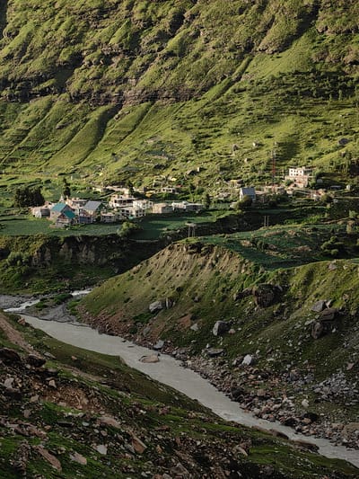 This is a high-angle, full shot of a village nestled in a steep, green mountain valley in Yari-Khoksar, India. A muddy river flows through the valley in the foreground, bordered by rocky, grassy slopes. Several houses, some with red-tiled roofs, are clustered together, surrounded by terraced fields. A telecommunications tower is visible near one of the houses. The mountain slopes are covered in lush green grass, with visible rock formations and patches of bare earth. The lighting suggests it is late afternoon, with sunlight casting long shadows. There are no people visible in the image.