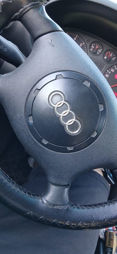 The image depicts an Audi steering wheel with the four-ring logo in the center. The steering wheel is black and shows signs of wear, particularly on the left side where the leather is damaged. The center of the steering wheel has an airbag cover labeled "AIRBAG." The dashboard and instrument panel are partially visible in the background, with illuminated gauges. A person is partially visible in the background wearing dark pants. A pack of cigarettes can be seen on the right side of the photo. There is no location in Tserovo, Bulgaria.
