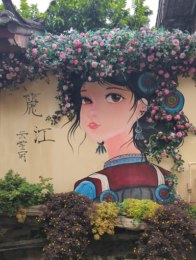 The image shows a mural of a woman on a tan-colored wall, located in Gucheng District, China. The mural depicts a woman with dark hair adorned with pink roses and green leaves. She has large, expressive brown eyes, pink lips, and wears a traditional outfit with blue and red accents. On the wall to the left of the mural, there are two lines of Chinese calligraphy. The mural is partially covered by an overhang with a dark, tiled roof and is decorated with trailing pink roses and green foliage that extends down the sides and over the woman's hair. In front of the wall, a low ledge is filled with various potted plants, including a dark leafy plant with small yellow flowers and a lighter green plant with yellow flowers. The overall scene suggests an outdoor decorative element, possibly in a courtyard or along a street in an area with traditional architecture.