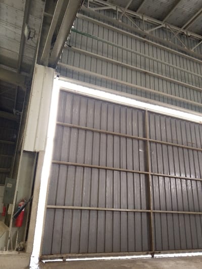 The image shows a large, metal, sliding door, mostly closed, inside a building with a concrete floor and a metal roof. The door is made of vertical panels, and a bright light is visible coming in from the gap between the door and the wall.  A red fire extinguisher sits on the floor near the wall to the left of the door.  The location is inside a building at latitude 22.694, longitude 90.601, in the city of Pangasia, Bangladesh.