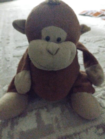 The media file shows a close-up, low-resolution image of a plush toy monkey sitting on a textured surface, likely a carpet or rug. The monkey is primarily brown with a lighter tan face, round black eyes, and a sewn-on nose and mouth. Its arms and legs are visible, positioned as if it is sitting upright. In the background, out of focus, several rectangular objects with orange and white colors are visible, possibly cards or other flat items. The lighting appears dim, suggesting an indoor setting, possibly during the evening or in a poorly lit room. The texture of the surface beneath the monkey is grainy and indistinct, with some lighter and darker patches creating a mottled effect. The overall impression is a candid, possibly playful, domestic scene. There is no visible text in the image. The location context provided is "Калыс-Ордо жаңы курулушу, Kyrgyzstan," suggesting the photo was taken in a new construction area within Kyrgyzstan.