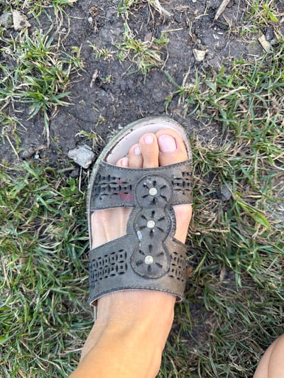 A close-up, top-down shot shows a person's left foot wearing a dark gray, open-toed sandal. The sandal has cut-out patterns and decorative circular embellishments with small, clear stones. The foot, with light skin and short, natural-colored toenails, is positioned on uneven ground covered with dry grass, dirt, and scattered small stones. The scene appears to be outdoors, possibly in a park or undeveloped area, consistent with the outskirts of a rural settlement like Petryaksa, Russia, where such terrain is common.