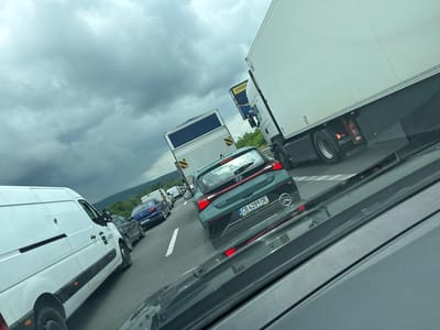 The image captures a traffic jam on a highway, likely in Bulgaria given the license plates. The scene is dominated by vehicles, including a white van with "SUNOTEC" branding, a dark blue Volkswagen, and a green Hyundai i20 with a license plate reading "CB 6289 OE". Behind these, several other cars and trucks are visible, indicating a significant traffic standstill. A large white semi-trailer truck with "SCANIA" visible on its side is also present, along with another truck with a blue and yellow sign. The sky is overcast with heavy, dark clouds, suggesting a possibility of rain or an impending storm. The time of day appears to be daytime, but the weather conditions create a somewhat somber atmosphere. The overall mood is one of frustration and waiting, typical of being stuck in traffic. The setting is a multi-lane highway surrounded by green hills in the distance.